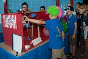 Students with red clown noses and neon colored wigs stand around contraption while judges look on.