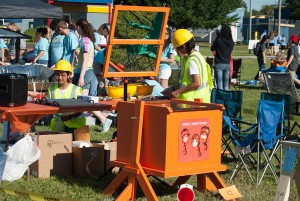 Girls in yellow hard hats and yellow construction vests prepare food to be cooked in their orange colored solar cooker.