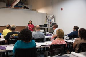 Instructor in front of seated students with an instructional model of a home in the background.