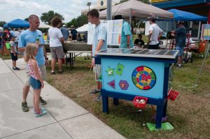 Father and daughter listen to Howard Middle School team member explain how project works. Solar panel on top of rectangular box, raised with legs, in which soft dart board and suction cup puzzle pieces are incorporated.