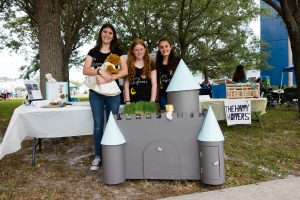 Three female students stand in front of their castle home designed for a cat.