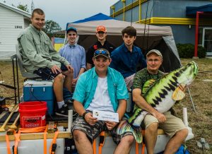 Five male students and instructor sits on their pontoon barge equipped with cooler, fishing poles and trolling motor.