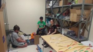 Three students sitting around contraption, which spanned from a long table and onto the floor.