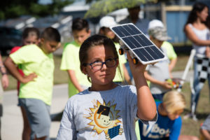 Student at EnergyWhiz event holding up Junior Solar Sprint model car in sun, partially shading face.