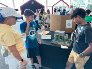 Adult talks to two-person student team about their Energy Inspired Art project called Eco House.
